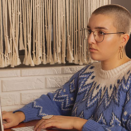 Mujer con pelo rapado y gafas trabaja frente a un portátil junto a una pared de macramé